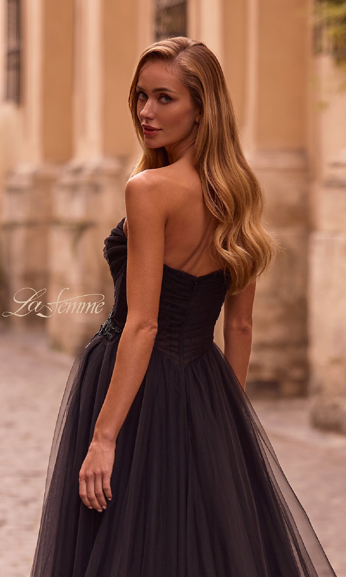 A woman in the La Femme 32789 strapless long prom ball gown with a sweetheart bodice and rhinestone floral detail stands outdoors, her long, wavy hair flowing as she glances over her shoulder.