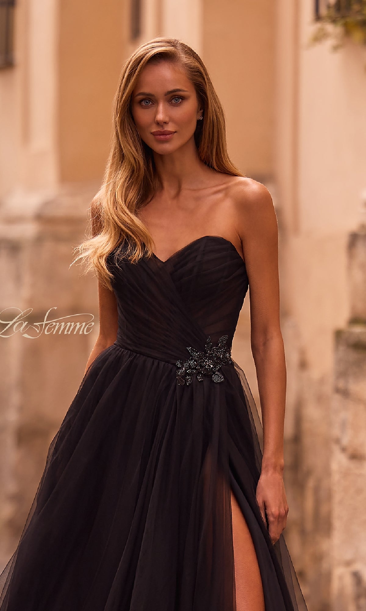 A woman with long blonde hair wears the La Femme 32789 strapless long prom ball gown with a sweetheart bodice, side slit, and rhinestone floral waist detail, standing outdoors.
