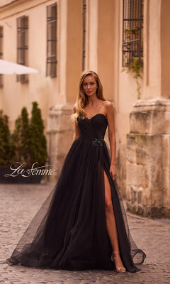 Wearing the Strapless Long Prom Ball Gown: La Femme 32789 with a sweetheart bodice and thigh-high slit, a woman stands on a cobblestone street beside an old building.