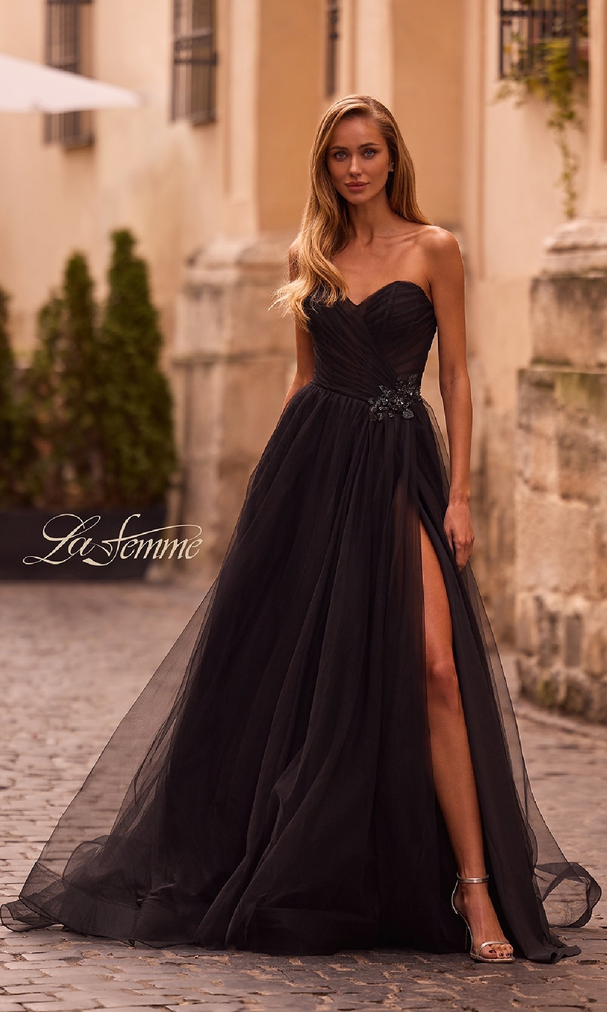 A woman stands outdoors in the black Strapless Long Prom Ball Gown La Femme 32789, featuring a sweetheart bodice, sheer overlay with a thigh-high slit, paired with silver high heels.
