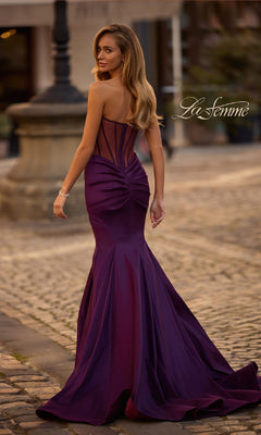 A woman in the Strapless Long Mermaid Prom Dress: La Femme 32791 stands on a cobblestone street, facing away, her fitted purple gown with a sheer back and elegant mermaid silhouette exuding sophistication.