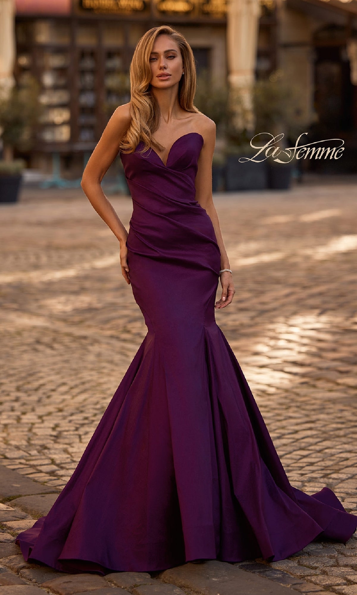 A woman in the Strapless Long Mermaid Prom Dress: La Femme 32791 stands outdoors on a cobblestone street, highlighting the elegance of this striking purple gown with 