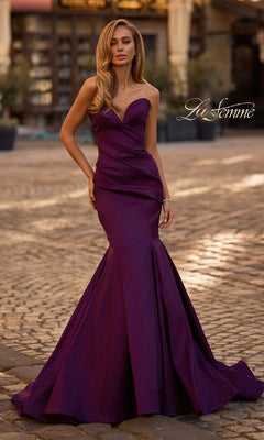 A woman in the Strapless Long Mermaid Prom Dress: La Femme 32791 stands outdoors on a cobblestone street, highlighting the elegance of this striking purple gown with "La Femme" displayed in the background.