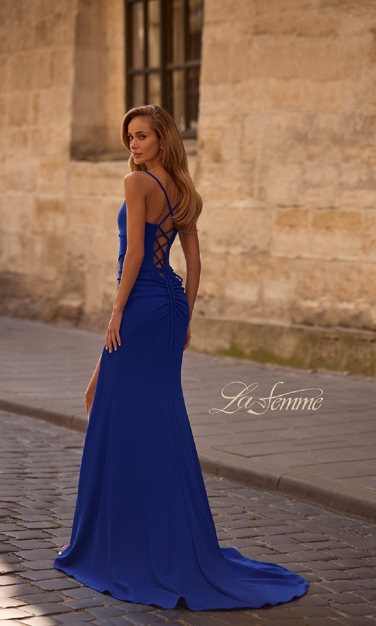 A woman in the floor-length La Femme 32802 prom dress, featuring a lace-up open back, stands on a cobblestone street and looks over her shoulder. 