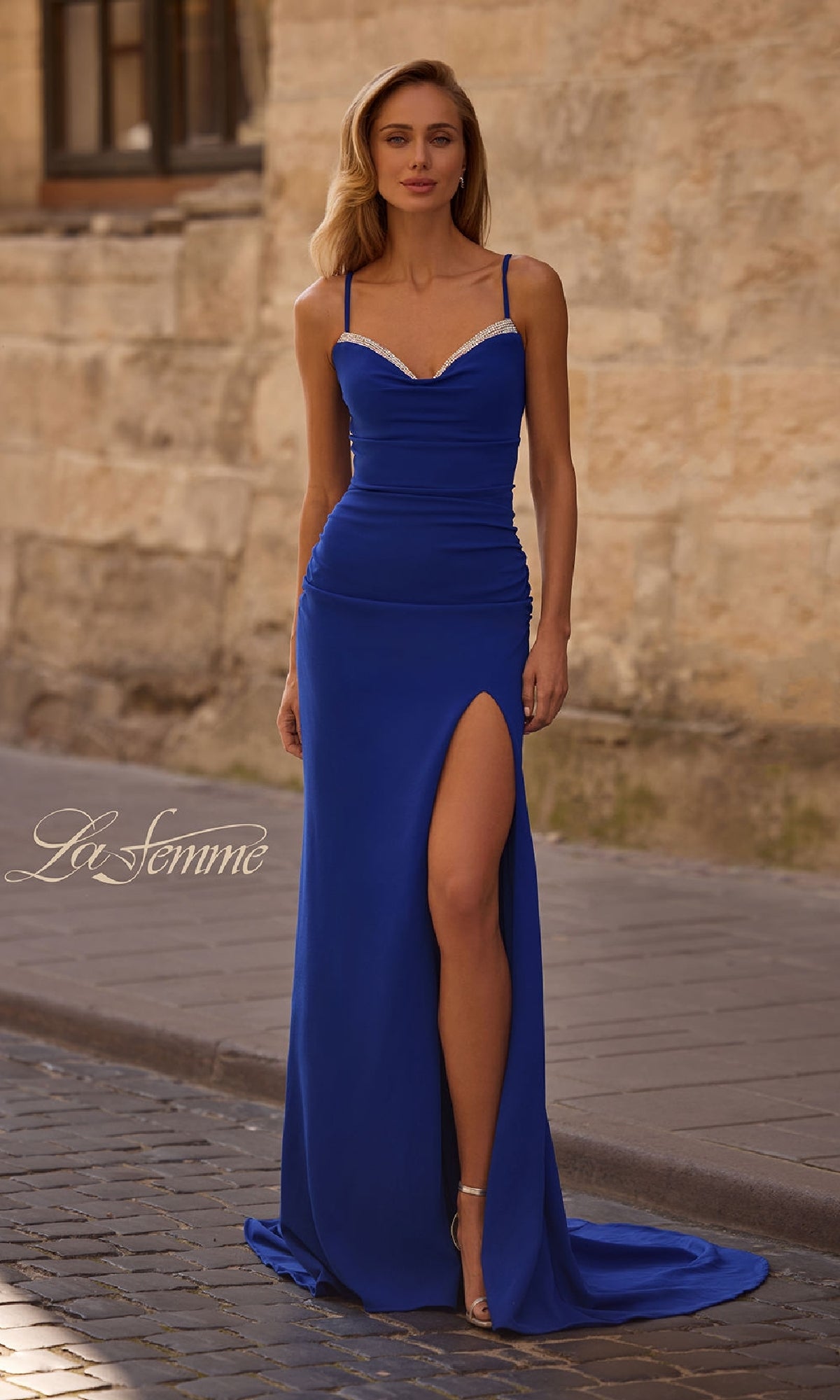 A woman in a fitted royal blue La Femme 32802 prom dress with spaghetti straps and a lace-up open back stands on a cobblestone street beside a stone wall.