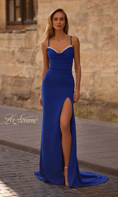 A woman in a fitted royal blue La Femme 32802 prom dress with spaghetti straps and a lace-up open back stands on a cobblestone street beside a stone wall.