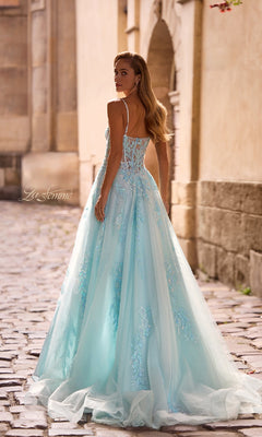 A woman in the La Femme 32810 long prom dress, featuring light blue tulle with beaded lace appliqué and a corset bodice, stands on a cobblestone street looking over her shoulder.