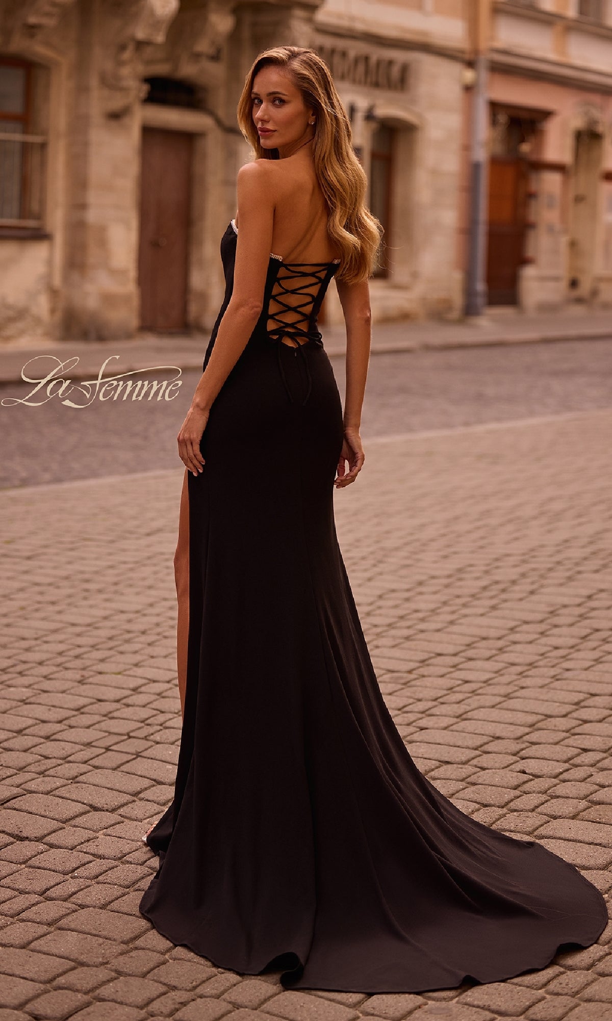 Wearing the La Femme 32829 Lace-Up Strapless Long Prom Dress with beaded trim, a woman stands on a cobblestone street and looks over her shoulder.