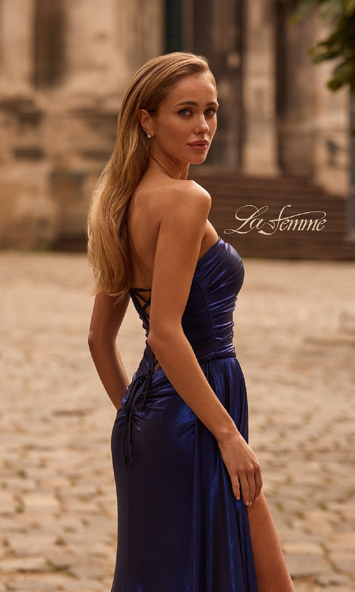 A woman stands outdoors on a cobblestone path, looking over her shoulder in the Strapless Long Metallic Prom Dress: La Femme 32830 in shiny dark blue, with the “La Femme” logo and La Femme 32830 visible in the background.