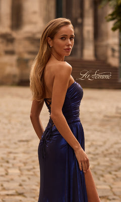 A woman stands outdoors on a cobblestone path, looking over her shoulder in the Strapless Long Metallic Prom Dress: La Femme 32830 in shiny dark blue, with the “La Femme” logo and La Femme 32830 visible in the background.
