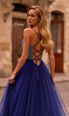 A woman in the La Femme 32838 cowl-neck long indigo prom dress stands outdoors, looking over her shoulder. The floor-length gown blends elegance and modern style with its stunning open back and crisscross straps.