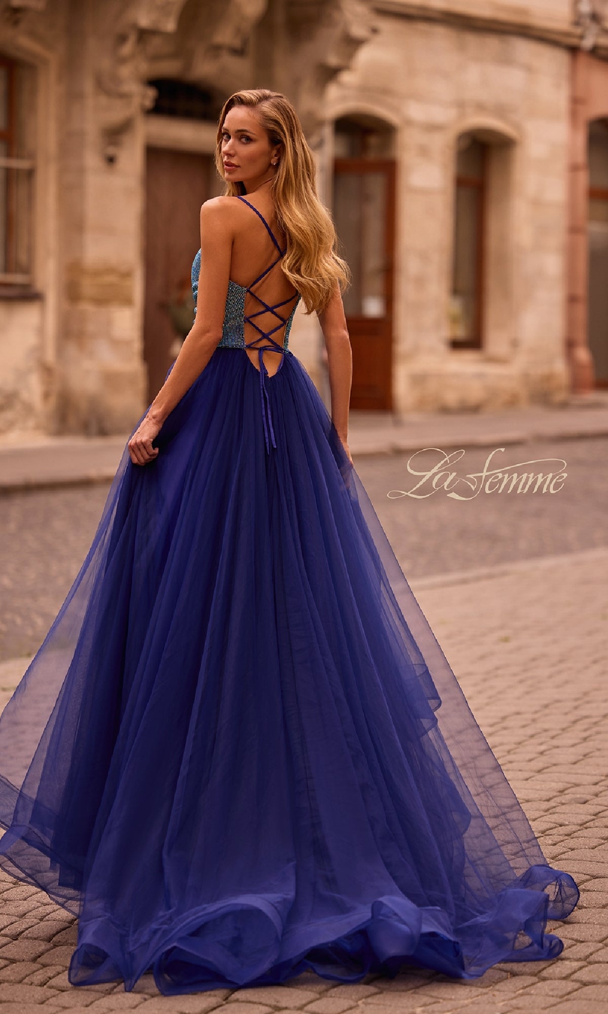 Woman in a Cowl-Neck Long Indigo Prom Dress (La Femme 32838) stands outdoors on a cobblestone street, facing away from the camera.