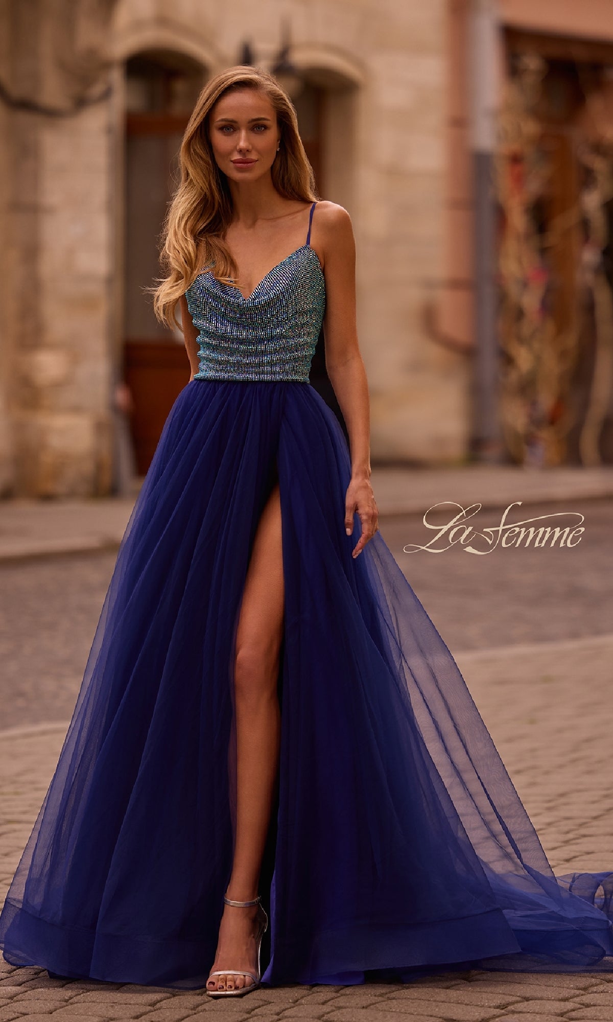 Wearing the La Femme 32838, a cowl-neck long indigo prom dress with a beaded bodice, thin straps, and high slit, she stuns on the cobblestone street.