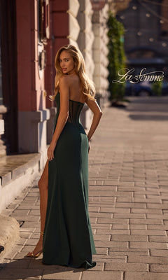 A woman in the La Femme 32849 Asymmetrical Strapless Prom Gown, a dark green long dress with a thigh-high slit, stands on a city sidewalk looking over her shoulder. "La Femme" appears in script on the right side.