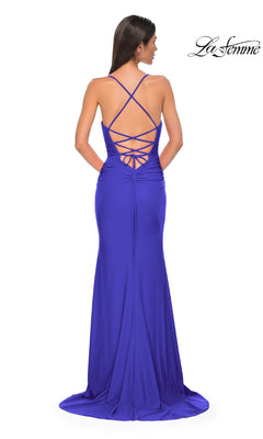 A woman stands facing away in a floor-length royal blue Strappy-Back Long Prom Dress: La Femme 32851, featuring a crisscross lace-up open back design.