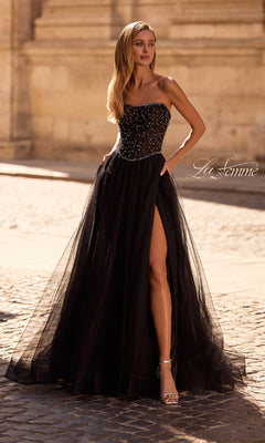 A woman in the La Femme 32856 Strapless Long Black Prom Ball Gown with a beaded bodice and thigh-high slit stands elegantly on a cobblestone street.