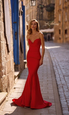 Wearing the Lace-Up Strapless Long Prom Dress: La Femme 32872, a woman stands on a cobblestone street by sandstone buildings and a blue door in daylight.