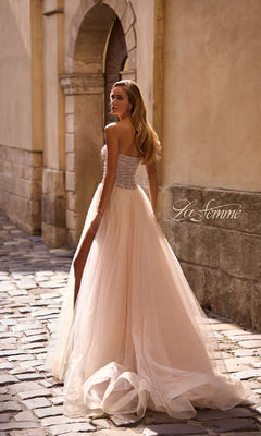 A woman wearing the Strapless Long Prom Ball Gown: La Femme 32875 with beaded details and a tulle skirt stands on a cobblestone street, glancing over her shoulder.