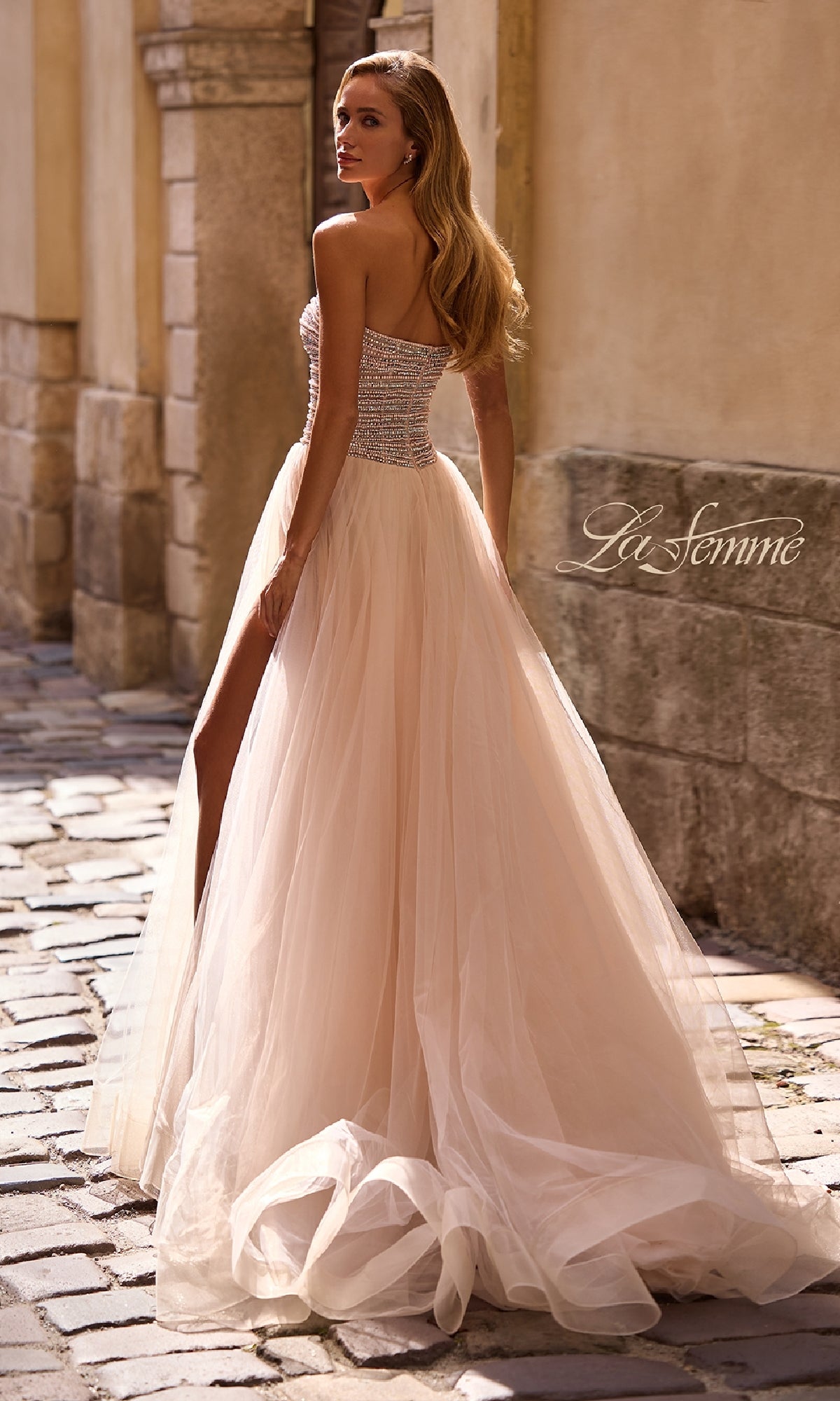 A woman wearing the La Femme 32875 Strapless Long Prom Ball Gown with beaded details and a high slit stands on a cobblestone street, glancing over her shoulder.