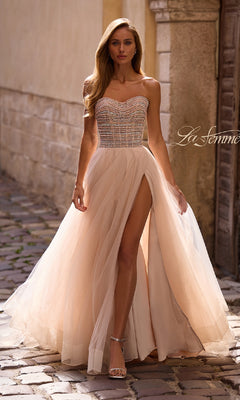A woman in the Strapless Long Prom Ball Gown: La Femme 32875, featuring beaded embellishments and a high slit, walks on a cobblestone street wearing strappy heels.