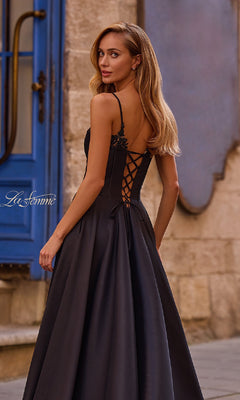 A woman in the La Femme 32878 Lace-Up Long Mikado Prom Dress, featuring a black formal design and lace-up back, stands outdoors on a stone street, gracefully looking over her shoulder.