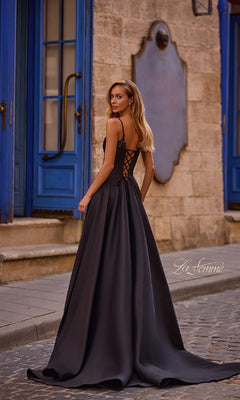Wearing the La Femme 32878 Lace-Up Long Mikado Prom Dress, a woman stands on a cobblestone street by blue doors and stone walls, looking over her shoulder.