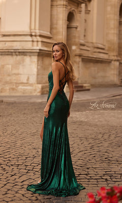 A woman in a long, metallic La Femme 32884 prom dress with lace-up details stands on a cobblestone street in front of a beige stone building.