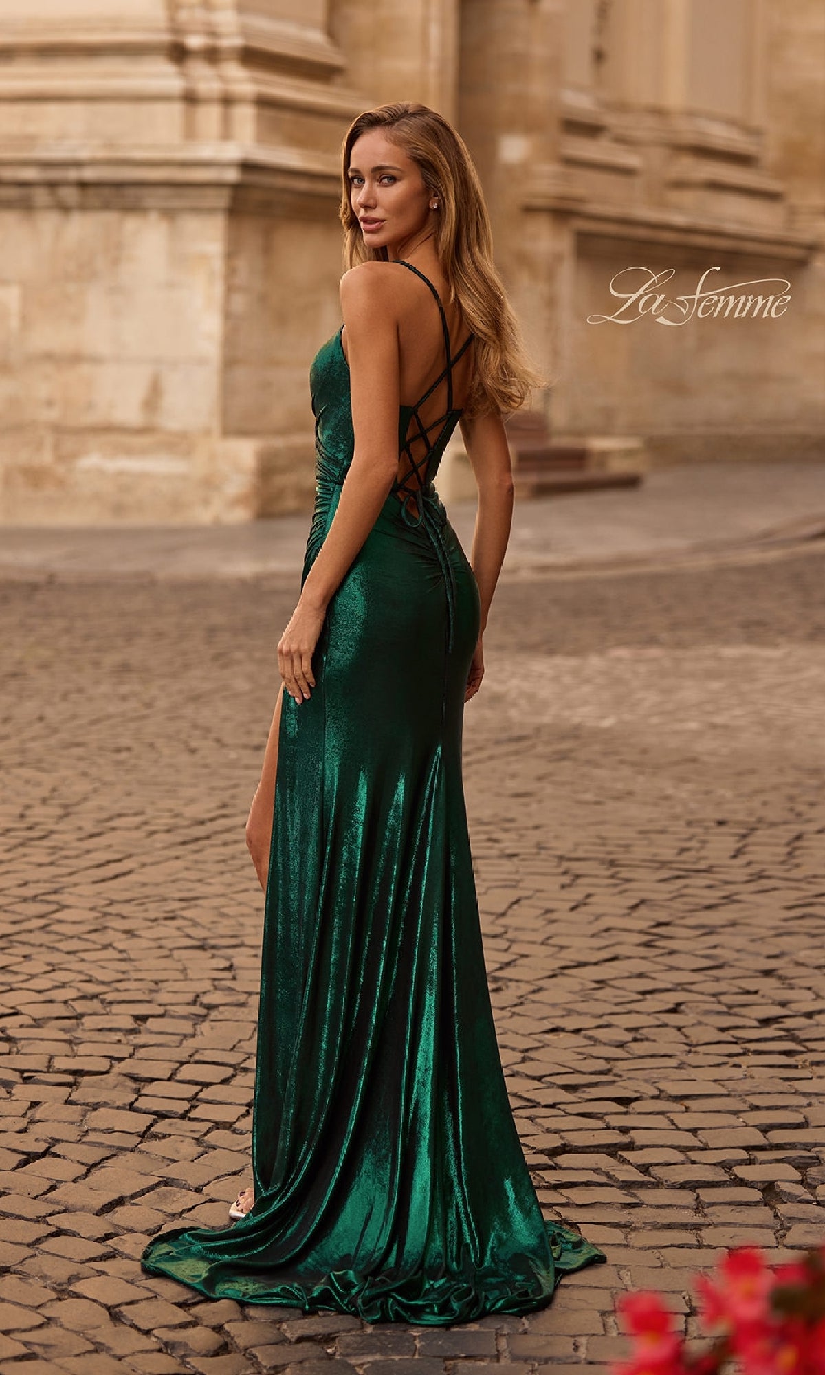 A woman in a long, dark green Lace-Up Long Metallic Prom Dress (La Femme 32884) with a high slit and crisscross back stands on a cobblestone street near a historic building.