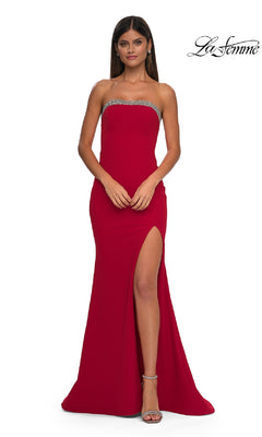 A woman wearing a red Bead-Trimmed Long Prom Dress: La Femme 32891 with a thigh-high slit and silver trim at the top, paired with silver high-heeled sandals, stands against a plain white background.
