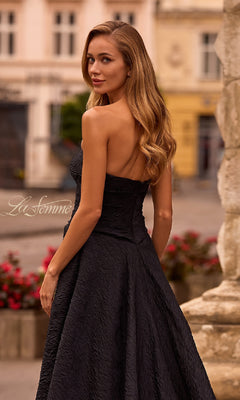 A woman with long wavy hair models the La Femme 32893 Textured Strapless Black Prom Dress outdoors near stone structures and flowers, glancing over her shoulder.