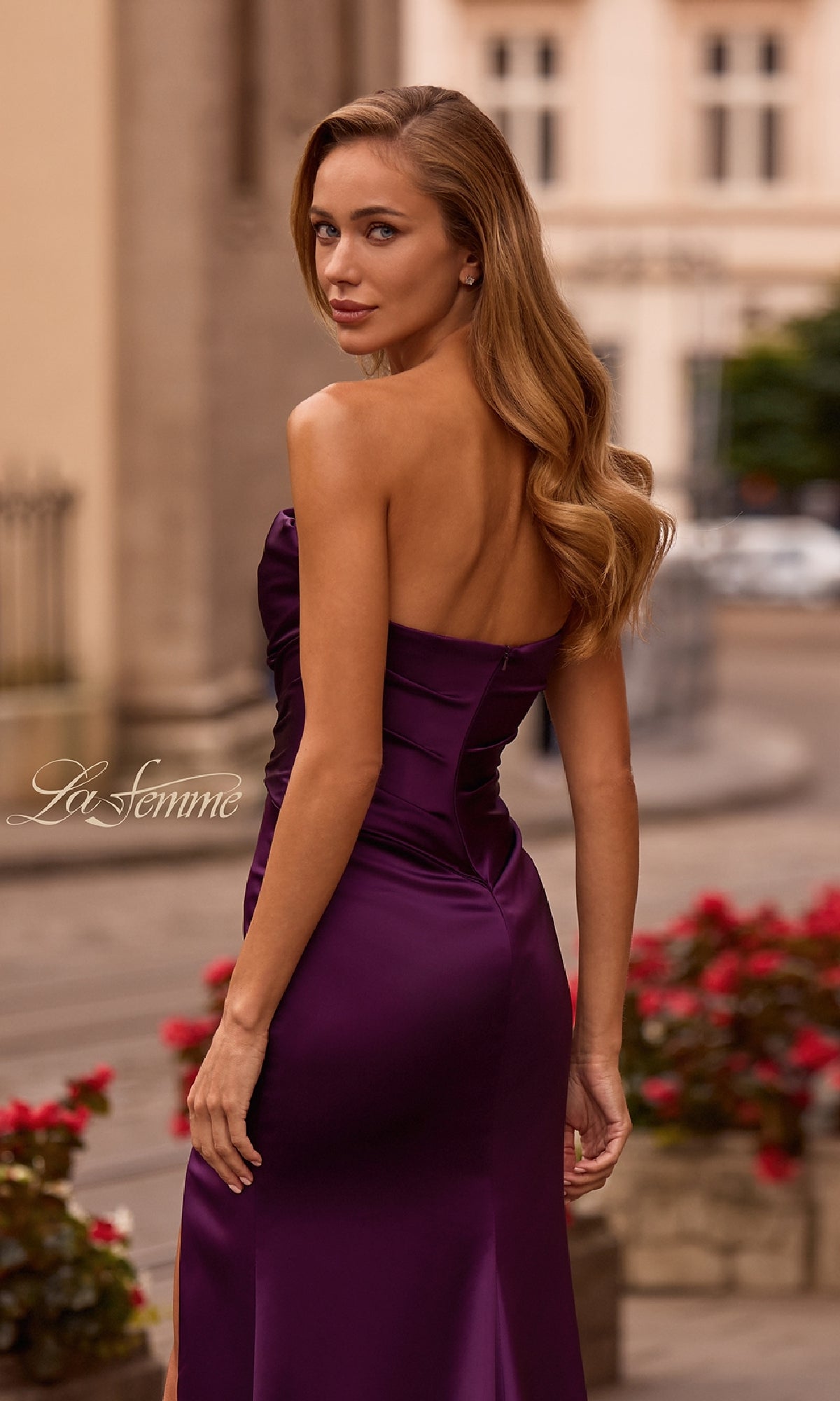A woman with long wavy hair wears the Strapless Sweetheart Prom Gown: La Femme 32896, a purple satin strapless prom dress, while standing outdoors by red flowers on a city street.
