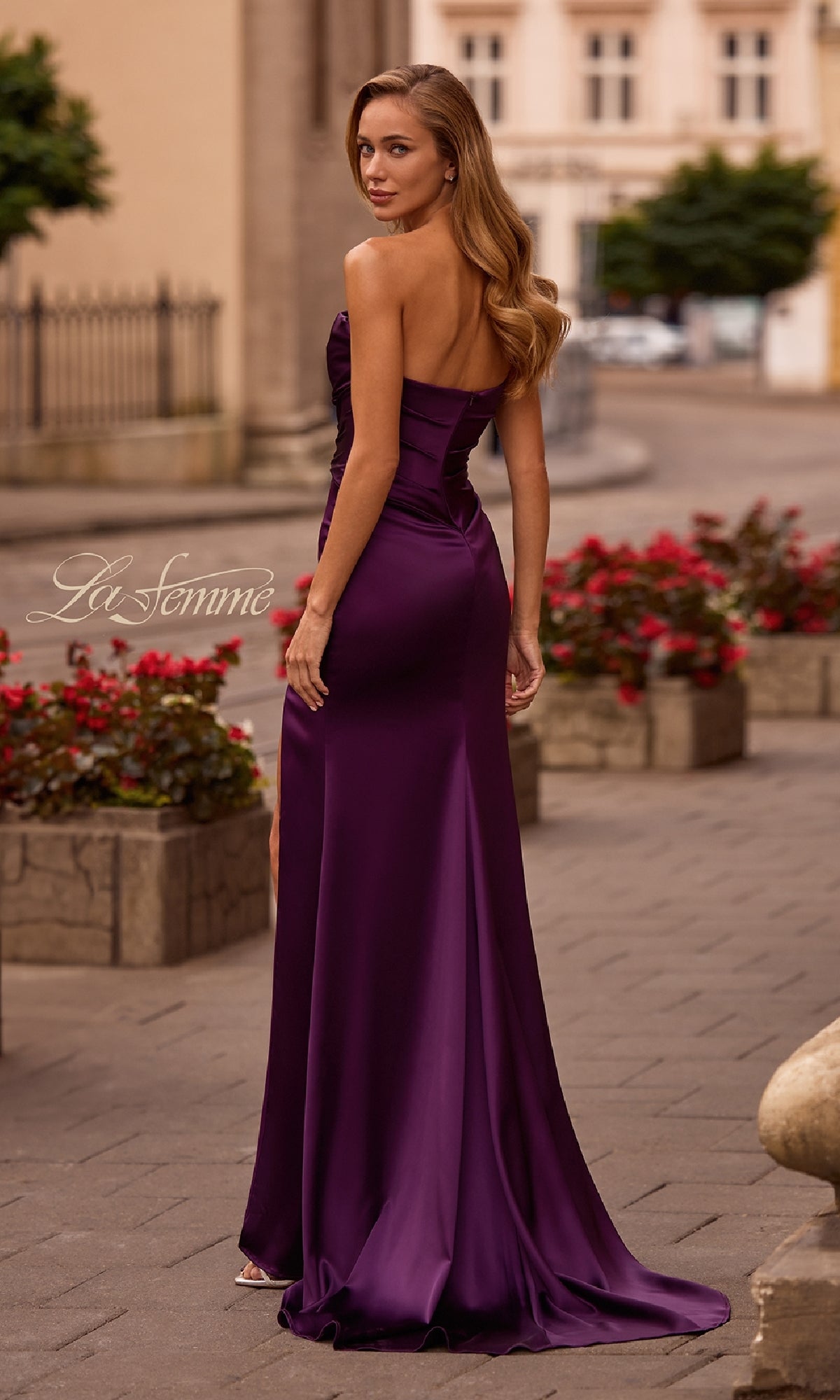 Wearing the Strapless Sweetheart Prom Gown: La Femme 32896, a woman stands outdoors on a paved walkway lined with red flowers and stone planters.
