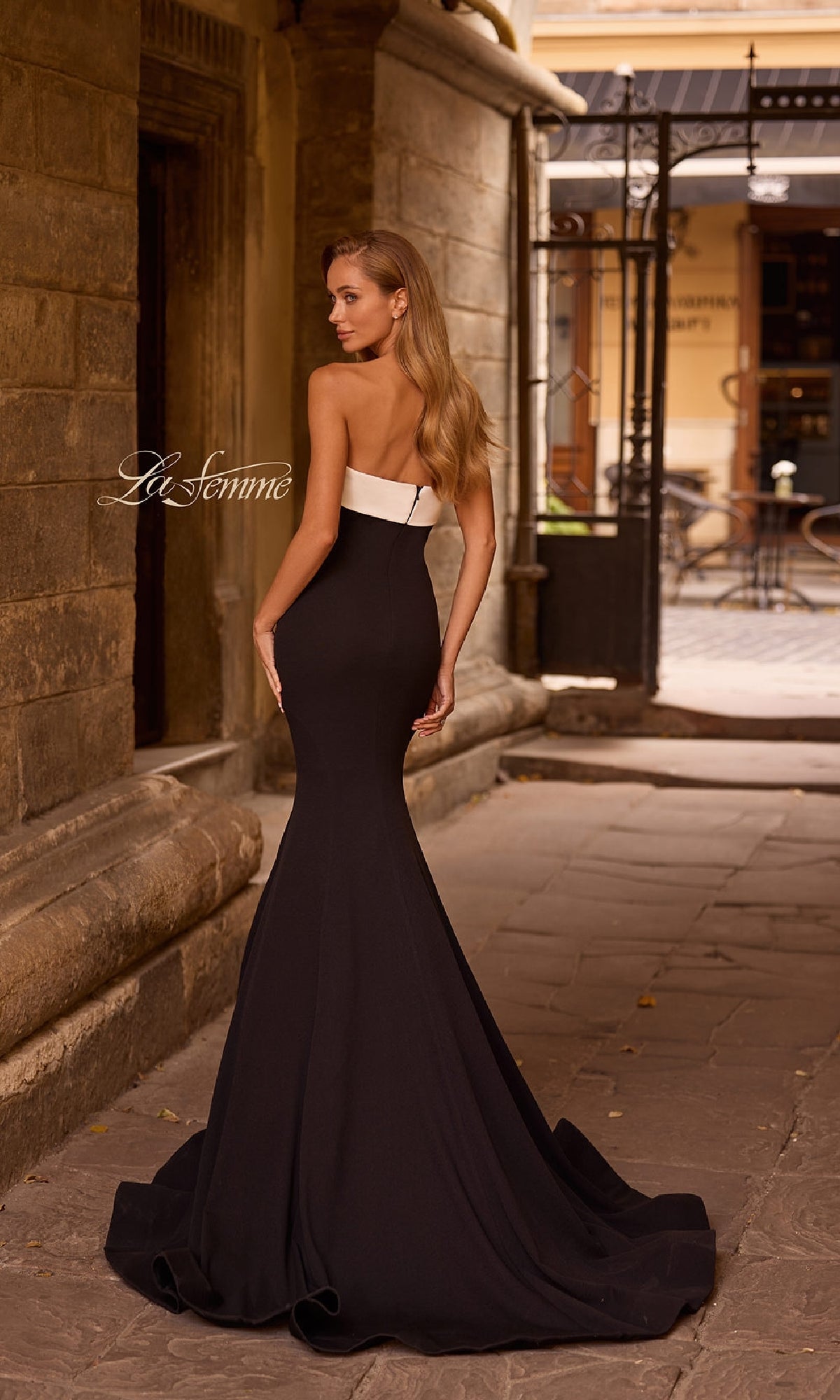 A woman in the La Femme 32914 strapless long black prom dress stands on a stone pathway in an old town, facing away from the camera.