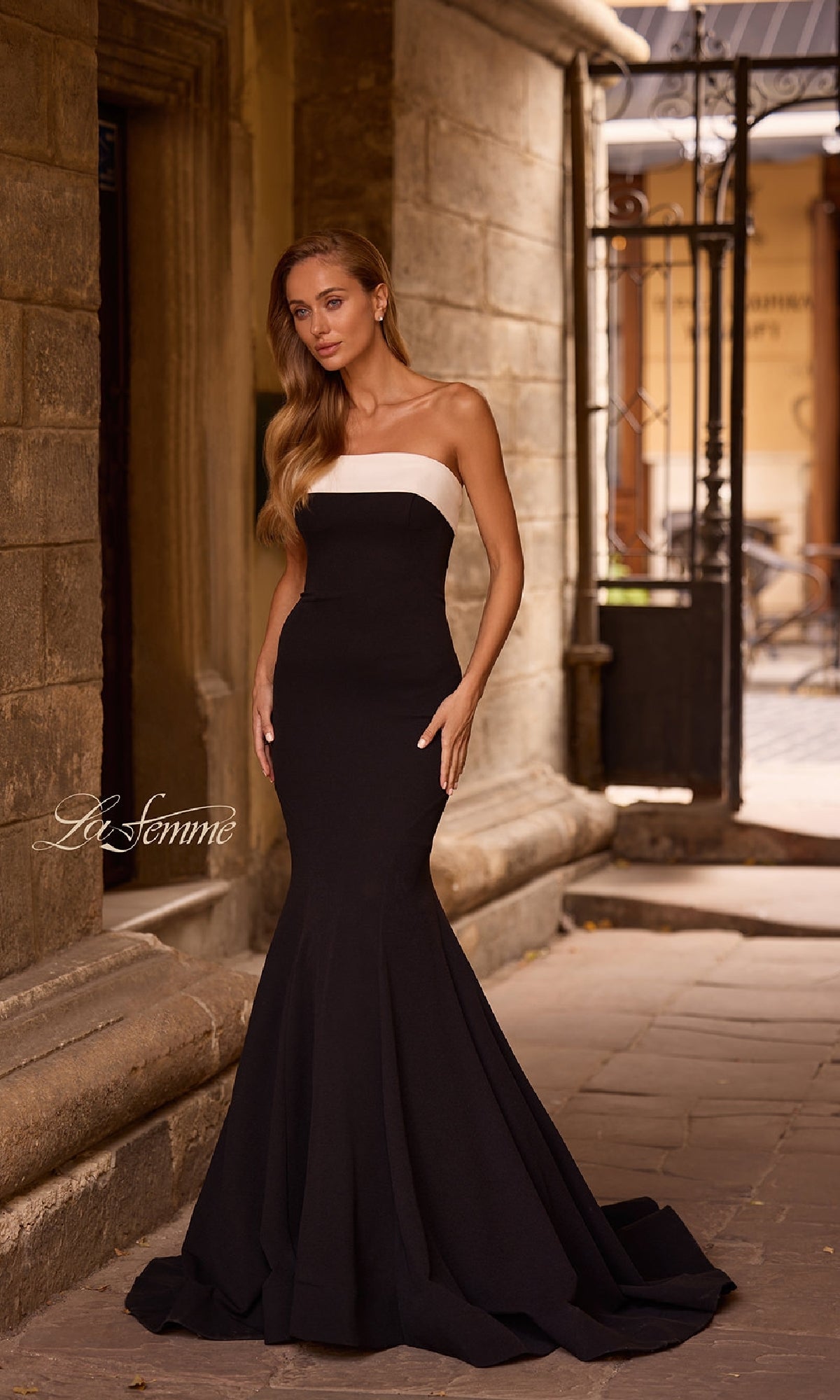 A woman wearing the La Femme 32914 strapless long black prom dress stands on a stone pathway near a historic building.