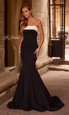 A woman in the La Femme 32914—strapless long black prom dress—stands on a stone walkway next to an old building.
