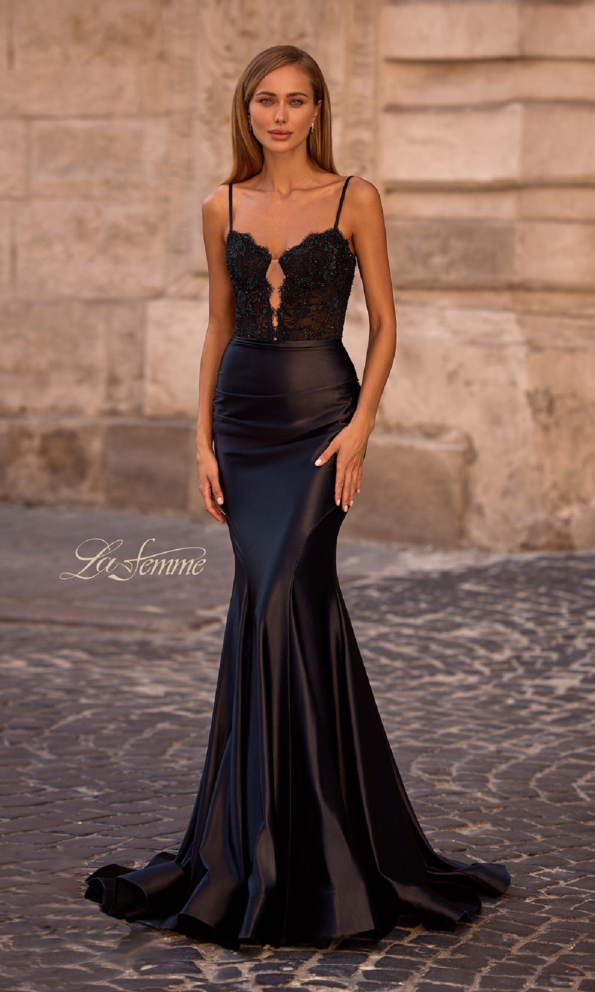A woman stands outdoors wearing the Black Mermaid Prom Dress: La Femme 32916, featuring a floor-length satin skirt, illusion lace bodice, and thin straps.