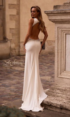 A woman stands outdoors on a cobblestone street beside a stone structure, wearing the Long Prom Dress: La Femme 32922 in white, featuring an open back and jeweled trim.