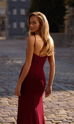 A woman in the Classic Long Sweetheart Prom Dress, La Femme 32925, featuring a fitted burgundy silhouette with a corset-style bodice, stands outdoors on a cobblestone street, looking over her shoulder.