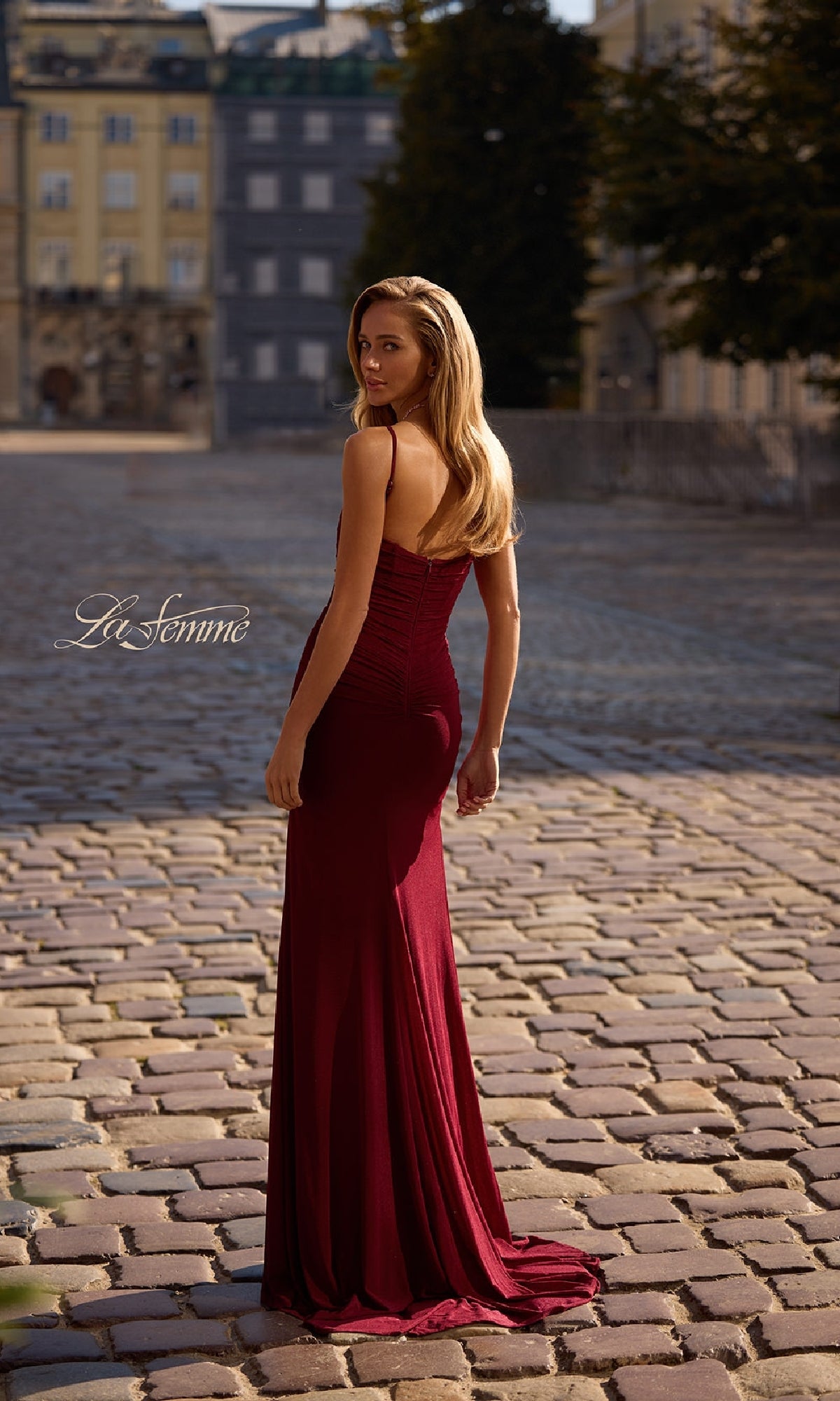 A woman in the La Femme 32925 Classic Long Sweetheart Prom Dress stands on a cobblestone street, glancing over her shoulder with buildings and trees in the background. The fitted burgundy gown features a corset-style bodice.