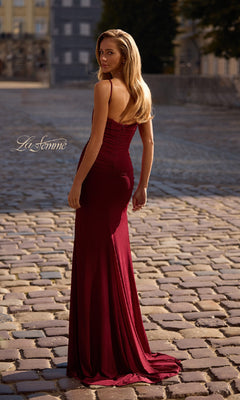 A woman in the Classic Long Sweetheart Prom Dress: La Femme 32925, a fitted, floor-length burgundy gown, stands on a cobblestone street facing away, glancing over her shoulder.