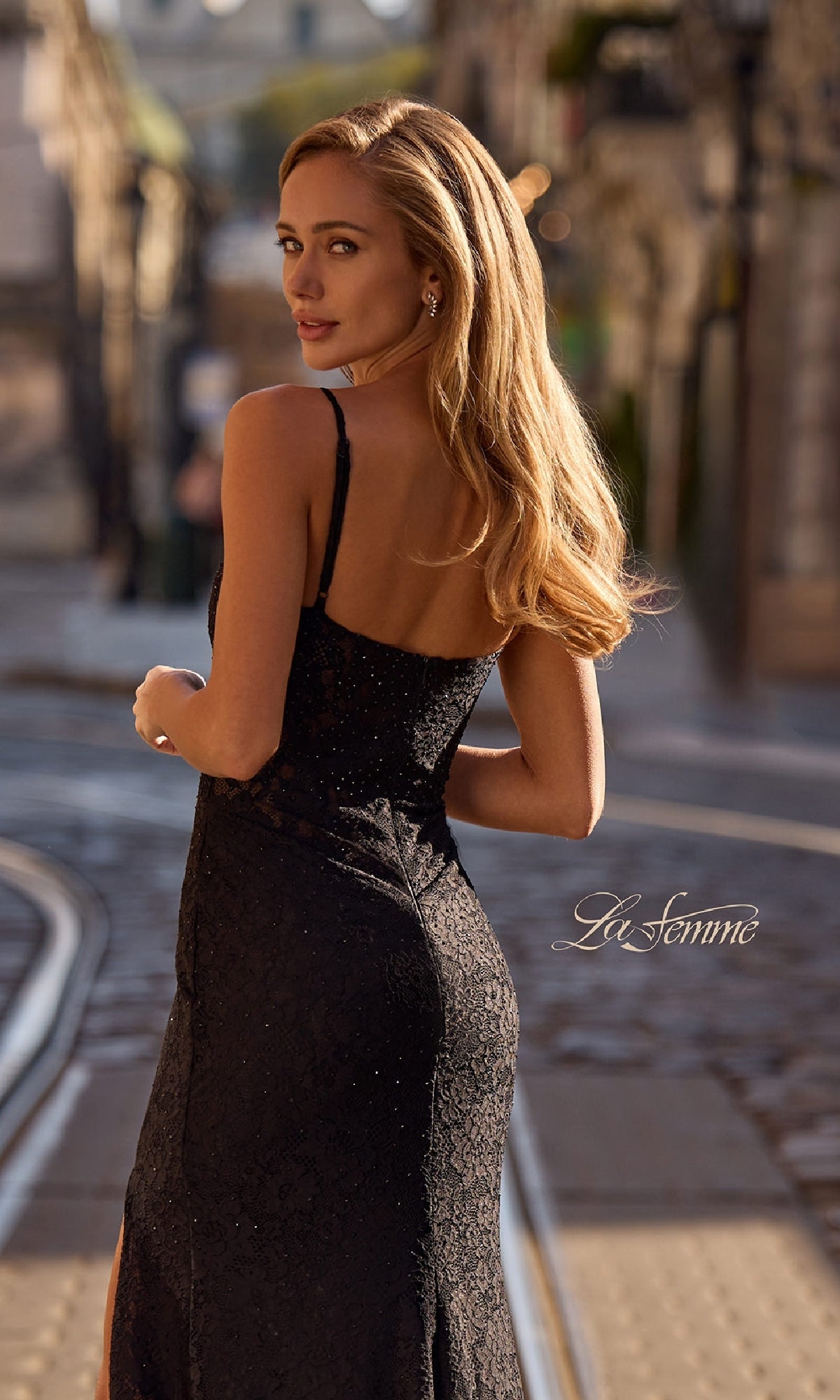 Woman with long blonde hair in the La Femme 32933 Long Beaded-Lace Prom Dress stands on a city street with tram tracks, glancing back over her shoulder.