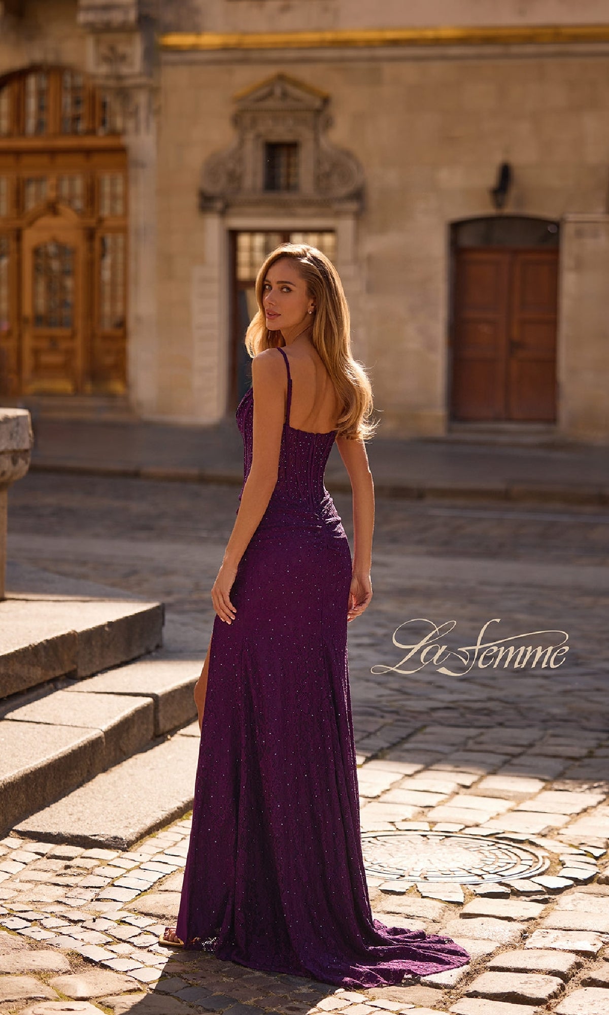 A woman wearing the Sheer-Waist Long Beaded Prom Dress: La Femme 32947 poses on a cobblestone street, glancing over her shoulder in an urban outdoor setting, with 