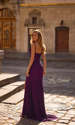 A woman wearing the Sheer-Waist Long Beaded Prom Dress: La Femme 32947 poses on a cobblestone street, glancing over her shoulder in an urban outdoor setting, with "La Femme" visible.