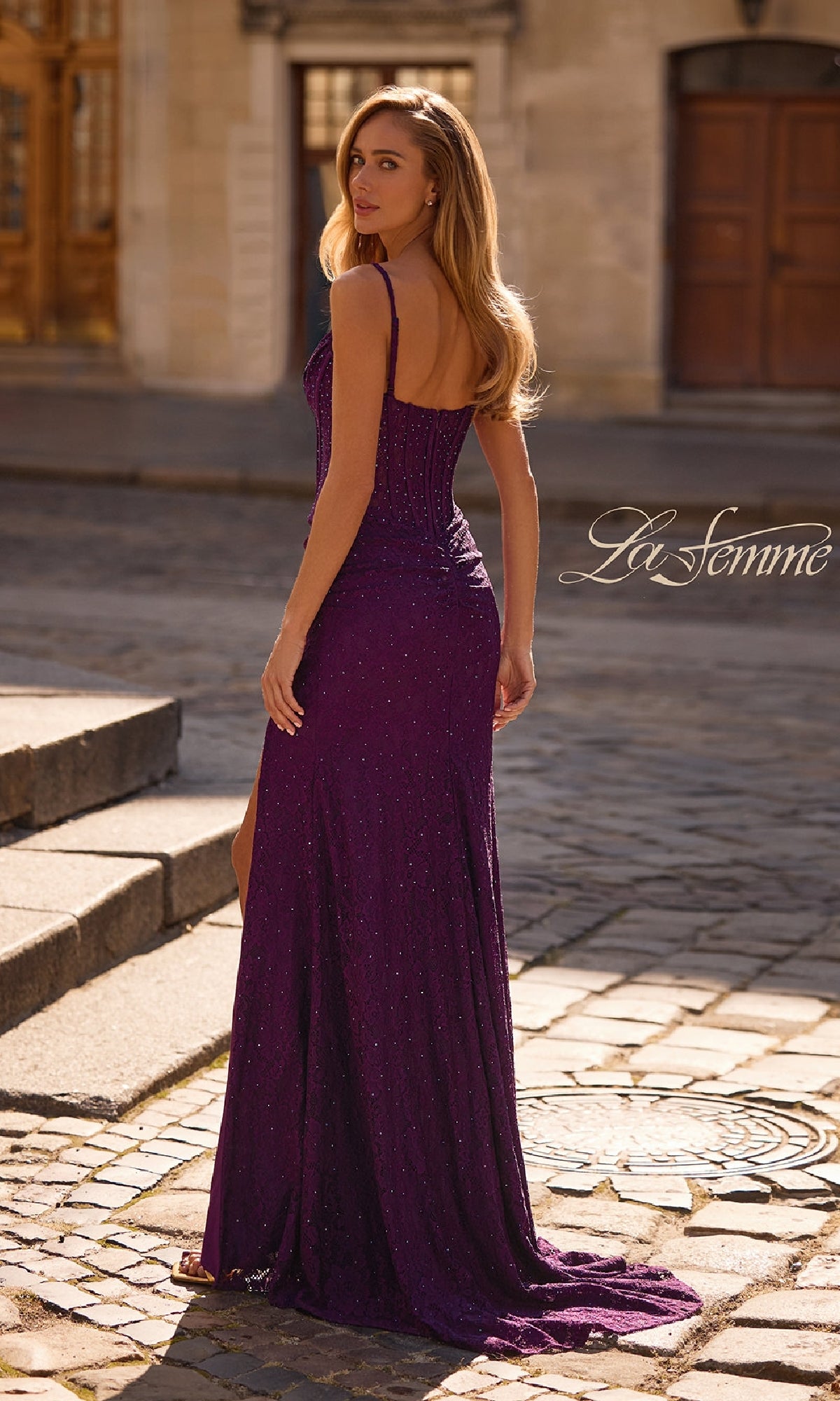 A woman in a fitted, purple Sheer-Waist Long Beaded Prom Dress (La Femme 32947) stands outdoors on a cobblestone street, looking over her shoulder and exuding elegance.