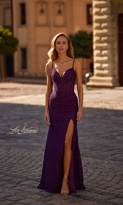 A woman in the Sheer-Waist Long Beaded Prom Dress (La Femme 32947) with sparkly purple details, spaghetti straps, and a thigh-high slit stands on a cobblestone street.