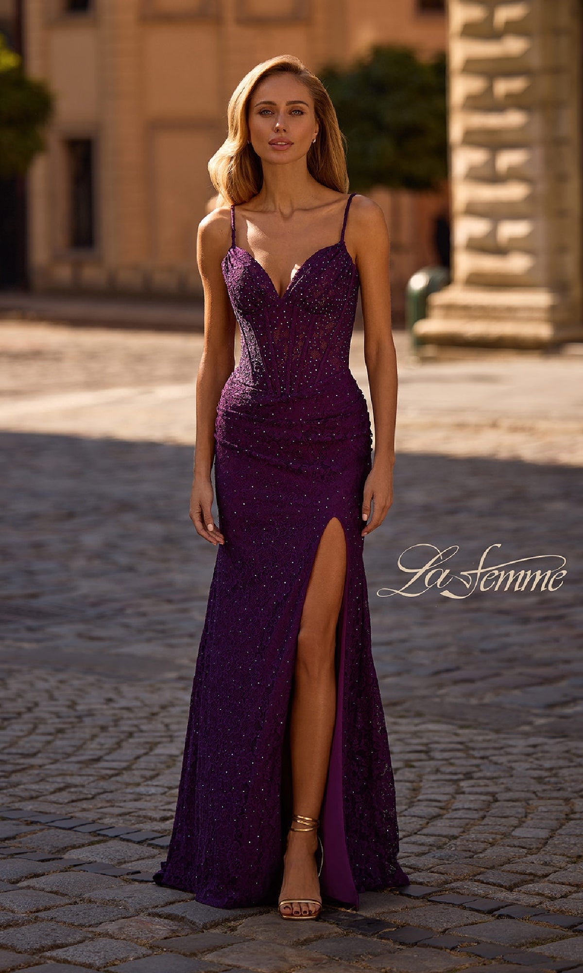 A woman stands outdoors in the Sheer-Waist Long Beaded Prom Dress (La Femme 32947), a fitted, floor-length purple gown with thin straps, a thigh-high slit, and sparkling beaded details. The 