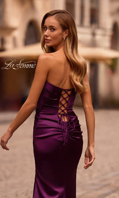 Woman in a deep purple Strapless Long Satin Prom Dress: La Femme 32952 with lace-up back stands on a cobblestone street, glancing over her shoulder.
