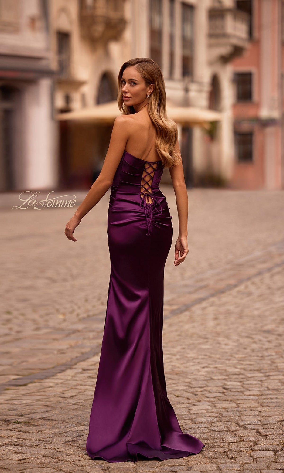 A woman wearing the La Femme 32952 strapless long satin prom dress with a lace-up back stands on a cobblestone street, looking over her shoulder.