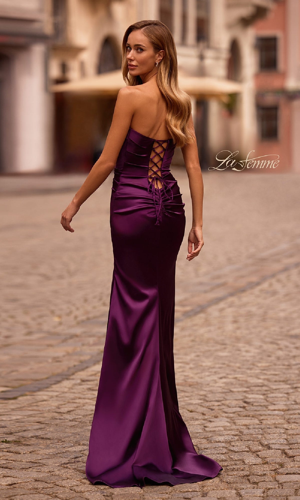 Wearing the La Femme 32952 Strapless Long Satin Prom Dress with a lace-up back, a woman stands on a cobblestone street, glancing over her shoulder.