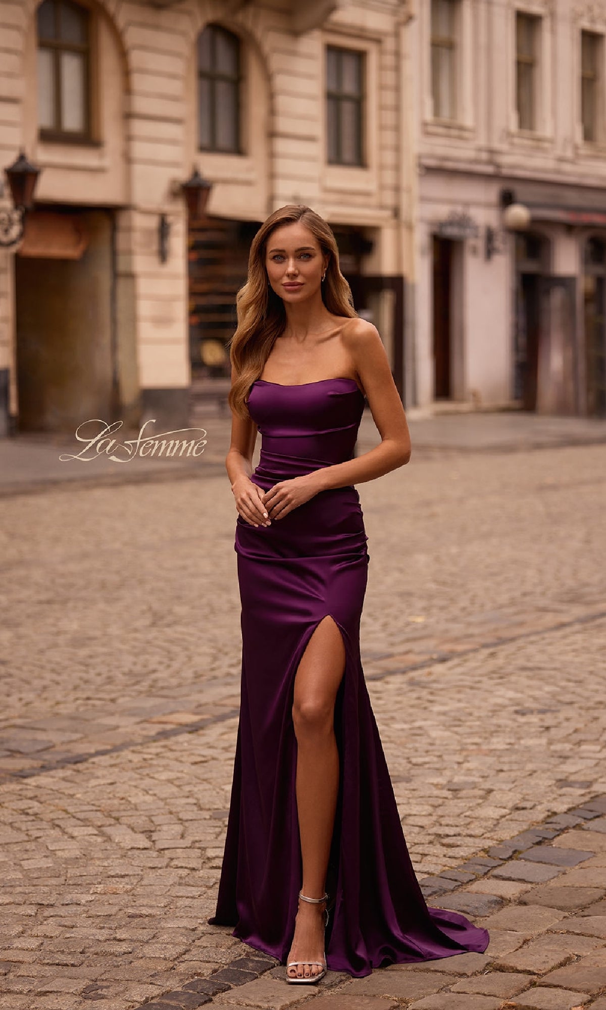 A woman wearing the Strapless Long Satin Prom Dress: La Femme 32952 with a thigh-high slit stands on a cobblestone street in front of historic buildings.